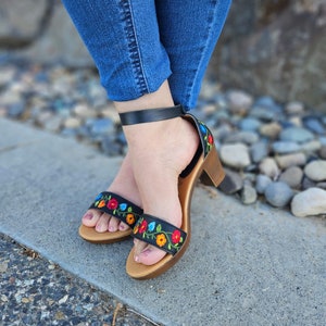 May include: Black heeled sandals with colorful embroidered flowers. The sandals have a black ankle strap and a wooden heel.