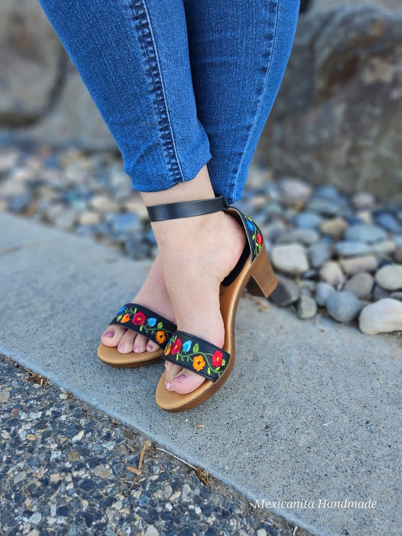 May include: Black heeled sandals with colorful embroidered flowers. The sandals have a black ankle strap and a wooden heel.