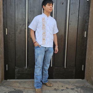 Puede incluir: Un hombre con una camisa blanca de botones con detalles bordados y pantalones vaqueros azules. Est&aacute; de pie frente a una puerta de madera.