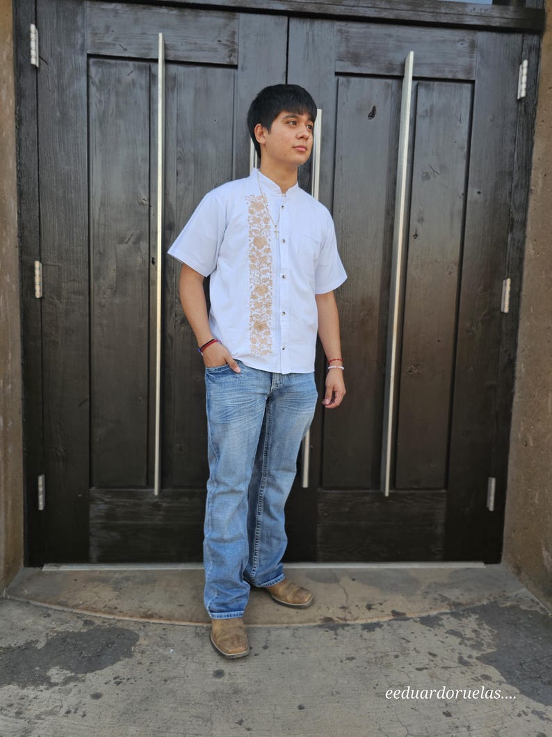 Puede incluir: Un hombre con una camisa blanca de botones con detalles bordados y pantalones vaqueros azules. Est&aacute; de pie frente a una puerta de madera.