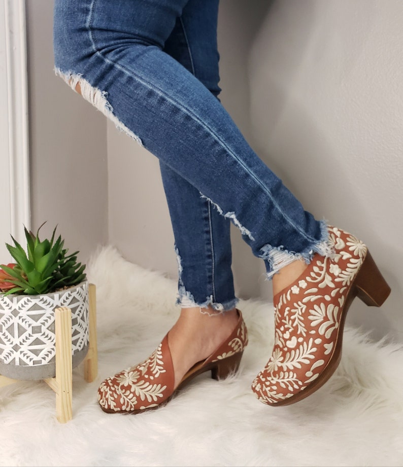 May include: Brown leather shoes with white floral embroidery. The shoes have a wooden heel and a cutout design. The person is wearing blue jeans with distressed details. The shoes are a unique and stylish footwear option.
