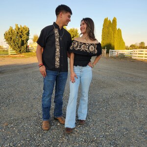 May include: A couple wearing traditional Mexican clothing. The man is wearing a black embroidered shirt with a white collar and blue jeans. The woman is wearing a black embroidered off-the-shoulder top with a white collar and blue jeans.