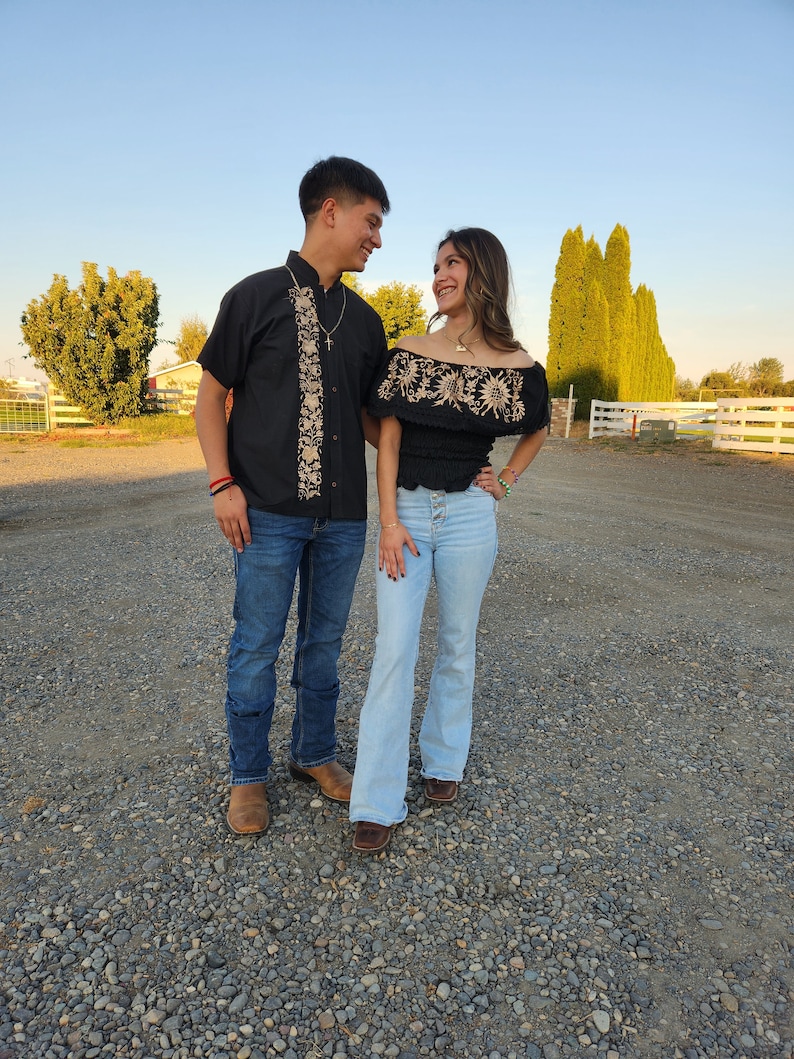 May include: A couple wearing traditional Mexican clothing. The man is wearing a black embroidered shirt with a white collar and blue jeans. The woman is wearing a black embroidered off-the-shoulder top with a white collar and blue jeans.