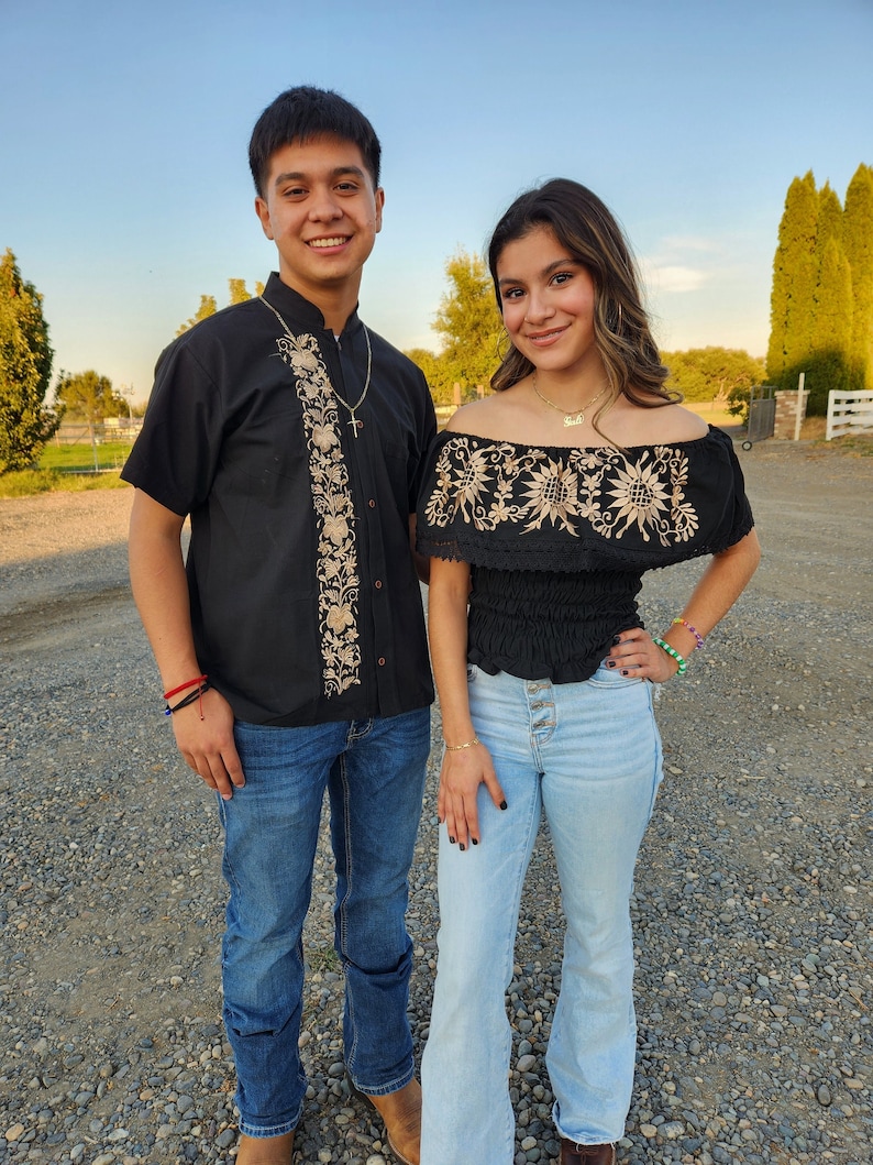 May include: A couple wearing traditional Mexican clothing. The man is wearing a black shirt with gold embroidery and blue jeans. The woman is wearing a black top with white embroidery and light blue jeans.