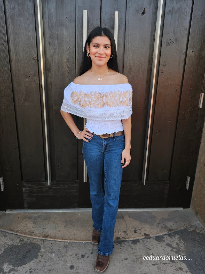 Puede incluir: Una mujer con un top blanco de hombros descubiertos con bordados dorados y jeans azules. Est&aacute; de pie frente a una puerta de madera.