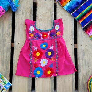Pode incluir: Um vestido rosa sem mangas com bordado floral colorido. O vestido tem um decote redondo e uma saia franzida.