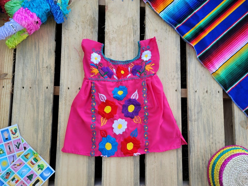 Pode incluir: Um vestido rosa sem mangas com bordado floral colorido. O vestido tem um decote redondo e uma saia franzida.