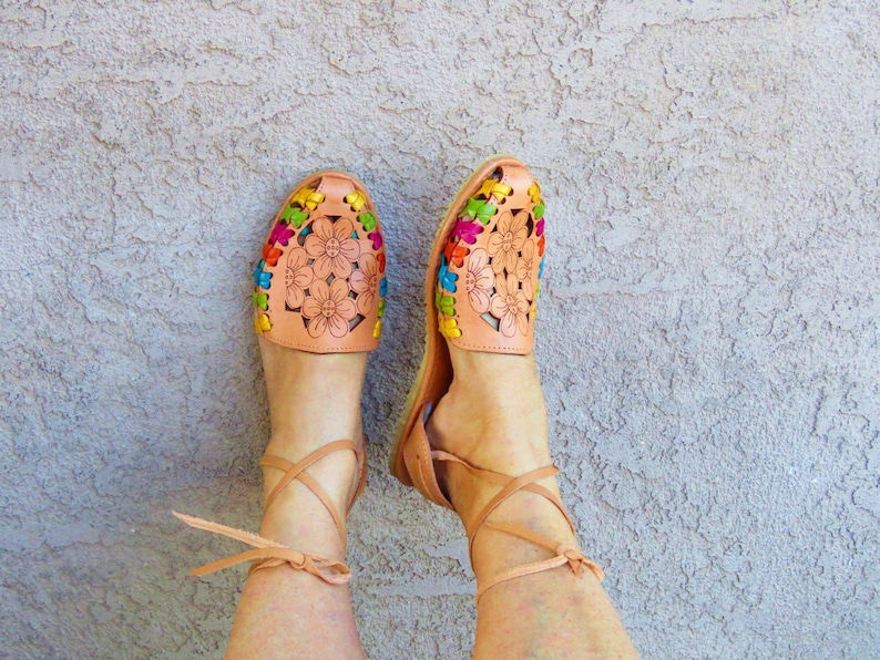 Puede incluir: Un par de sandalias de cuero marr&oacute;n con bordados florales de colores. Las sandalias tienen cordones que se atan alrededor de los tobillos.