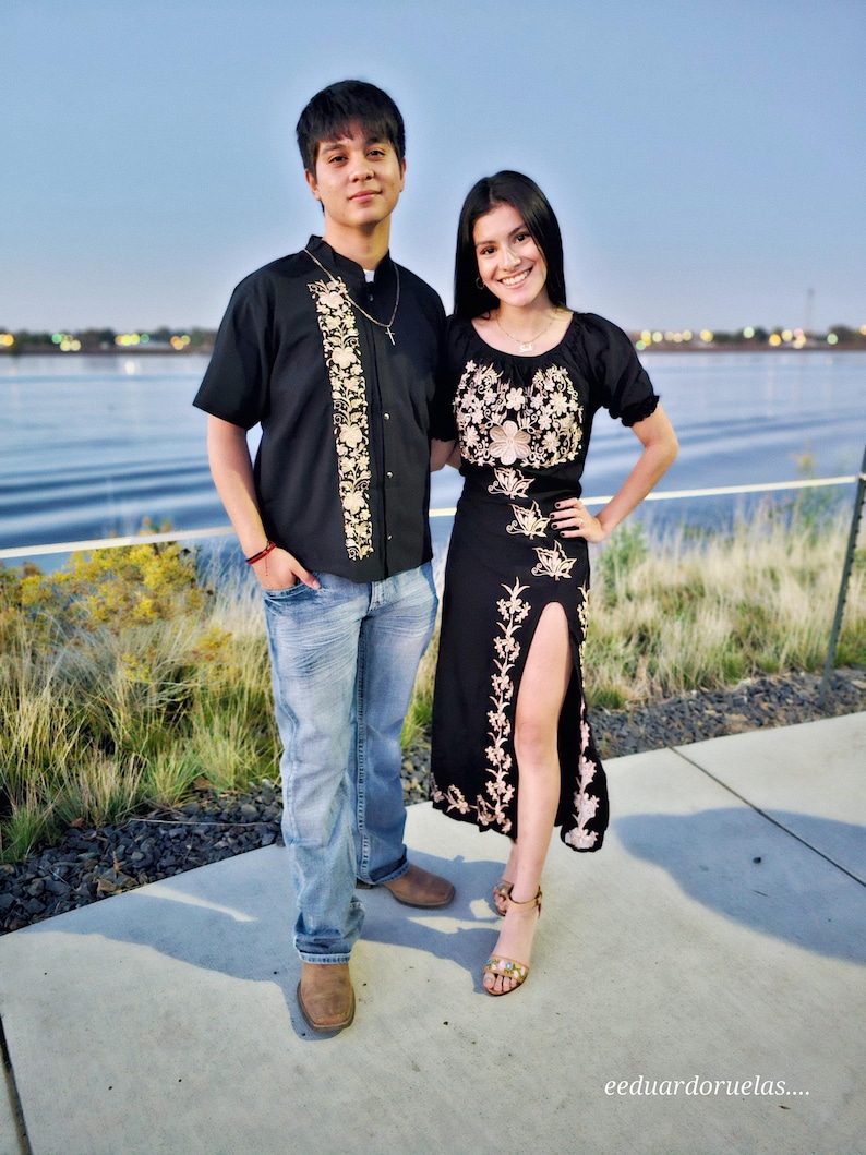 May include: A couple poses for a photo in front of a body of water. The man is wearing a black button-down shirt with gold embroidery and blue jeans. The woman is wearing a black dress with gold embroidery and a side slit.