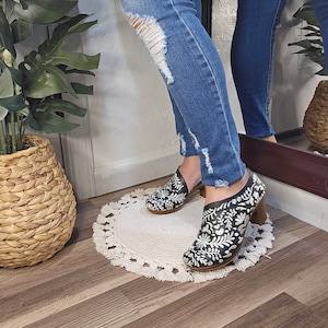 May include: Black leather ankle boots with white floral embroidery and a wooden heel. The boots are worn with blue denim jeans. The text "Mexicanita Handmade" is visible at the bottom of the image.
