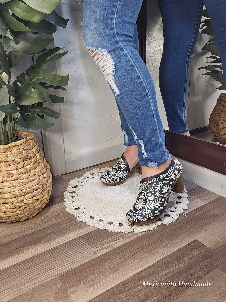 May include: Black leather ankle boots with white floral embroidery and a wooden heel. The boots are worn with blue denim jeans. The text "Mexicanita Handmade" is visible at the bottom of the image.