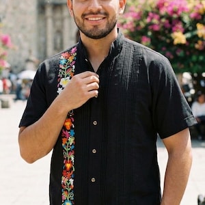 May include: A black short-sleeved shirt featuring a mandarin collar, a vertical row of buttons, and a prominent vertical panel of colorful floral embroidery in shades of red, orange, yellow, blue, and green along the right side of the chest.