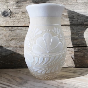Può includere: Un vaso in ceramica color crema con motivi floreali bianchi in rilievo. Il vaso ha una base larga, un corpo leggermente affusolato e un bordo svasato. I disegni floreali includono fiori e foglie stilizzati. Il vaso è esposto su una superficie di legno.