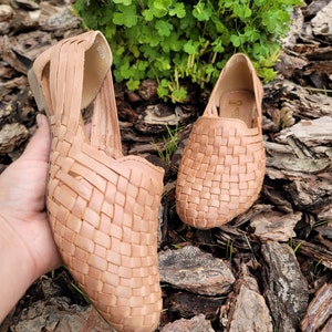 May include: A pair of woven leather sandals in a light brown color. The sandals have a woven design and are sitting on a bed of wood chips.