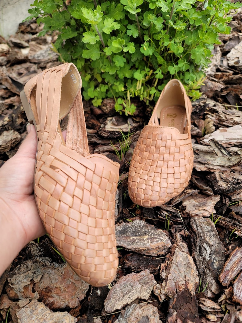 May include: A pair of woven leather sandals in a light brown color. The sandals have a woven design and are sitting on a bed of wood chips.
