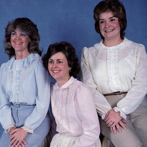 May include: Three women wearing white blouses with lace trim. The woman on the left is wearing a light blue blouse, the woman in the middle is wearing a pink blouse, and the woman on the right is wearing a white blouse. All three women are smiling.