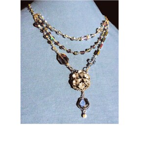 May include: A multi-layered necklace with a large, silver, crystal-encrusted pendant. The necklace is made of small, iridescent beads in shades of blue, purple, and gold.