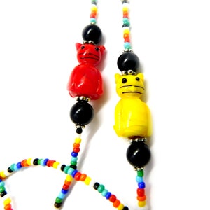 May include: A colorful beaded necklace featuring red and yellow cat-shaped beads, black beads, and a variety of smaller, multicolored beads. The cat beads are separated by black beads and silver-colored accents. The necklace has a playful and whimsical design.