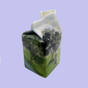 May include: A ceramic milk carton painted with a landscape scene of a green field, trees, and a person walking. The carton is white with blue and green accents.