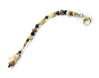 May include: A beaded charm featuring a small white ceramic animal figure with pink accents, strung with alternating cream-colored oval beads, dark brown spherical beads, and patterned round beads, finished with a gold-tone metal lobster clasp.