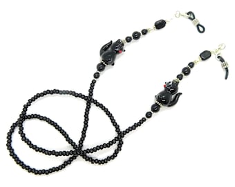May include: A black beaded eyeglass chain featuring two glossy black cat charms with small red accents. The chain is constructed with uniform round black beads, silver-toned spacers, and black rubber loops at each end for securing to eyewear frames.