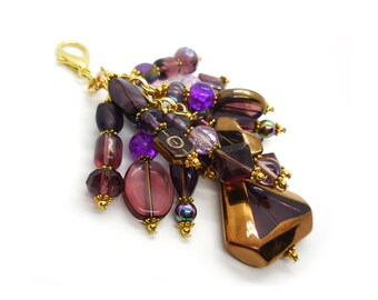 May include: A purple and gold beaded keychain with a large, faceted, purple and gold charm. The charm is shaped like a teardrop and has a metallic finish.