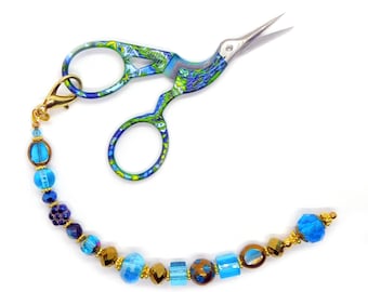 May include: Stork-shaped embroidery scissors featuring a vibrant blue and green patterned finish on the handles and blades. A gold-toned lobster clasp attaches a decorative beaded chain consisting of various blue, gold, and dark glass beads.