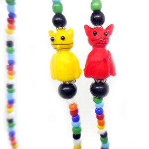 May include: A beaded necklace featuring two cat-shaped glass beads in bright yellow and red, accented with black spherical beads and smaller multicolored glass beads arranged in a long, continuous strand against a plain white background.