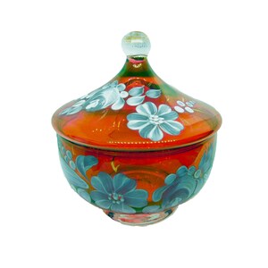 May include: A round, orange glass bowl with a matching lid, featuring a hand-painted floral pattern in light blue and white. The lid is topped with a clear glass knob, and the entire piece has a translucent, iridescent finish throughout.