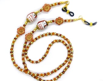 May include: Eyeglass chain with a beaded design in shades of gold, brown, and orange. The chain features two owl-shaped beads and flower-shaped accents. The ends have loops to attach to glasses.