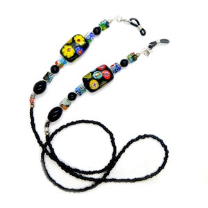 May include: An eyeglass chain featuring a long strand of small black beads, accented with larger black oval beads, rectangular black beads with colorful floral millefiori patterns, and small square multicolored glass beads, ending in black rubber loops.
