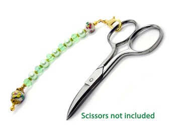 May include: A decorative scissor fob with a gold-colored clasp. The fob features a strand of light green beads, white pearls, and decorative floral beads. The text "Scissors not included" is visible in green.