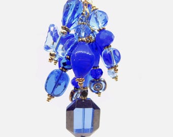 May include: A cluster pendant featuring various shades of blue glass beads in different shapes, including round, faceted, and butterfly-shaped beads. The beads are connected by small gold-toned metal spacers and links, creating a dense, dangling arrangement.
