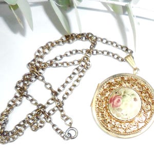 May include: A gold locket necklace with a delicate chain. The locket features a round, ornate design with a painted floral pattern in shades of pink, green, and white.