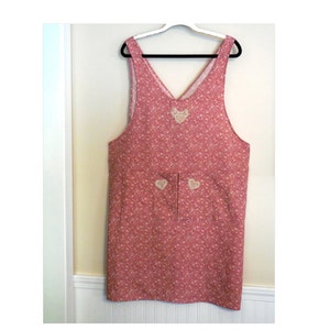 May include: A pink and white floral print apron with a V-neck and two pockets. The apron has white heart-shaped embroidery on the front and pockets.