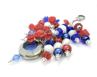 May include: A red, white, and blue beaded charm with a silver metal charm in the center. The charm is made of various beads, including red and white pearls, blue and red glass beads, and red plastic flowers.