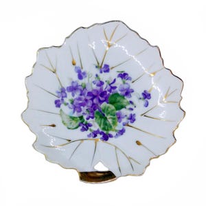 Puede incluir: Plato blanco de porcelana con forma de hoja, con borde dorado y detalles dorados. El plato presenta un ramo pintado a mano de violetas moradas y hojas verdes en el centro. Una pieza decorativa para servir o exhibir.