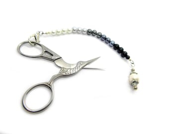 May include: Silver-toned metal embroidery scissors shaped like a bird with engraved wing details. A beaded chain is attached to the handle, featuring a gradient of white, grey, and black beads, ending with a single larger cream-colored bead.