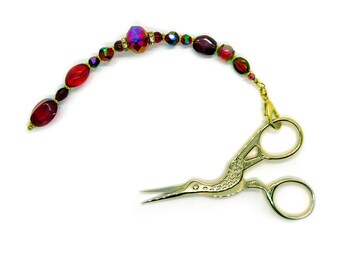 May include: Gold-toned stork-shaped embroidery scissors attached to a decorative beaded chain. The chain features a series of deep red, translucent, and faceted beads interspersed with small gold-colored metal spacers and accents.