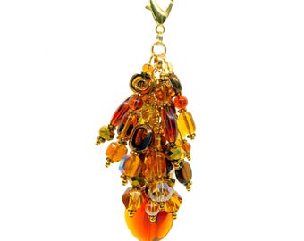 May include: A decorative charm featuring a cluster of amber, orange, and brown glass beads in various shapes and sizes, suspended from a gold-toned metal lobster claw clasp. The beads include faceted, round, and oval designs with metallic accents.