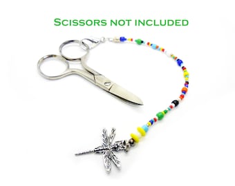 May include: A silver dragonfly charm attached to a beaded lanyard with colorful beads. The lanyard is connected to a pair of small, silver scissors. Text in green reads "Scissors not included."