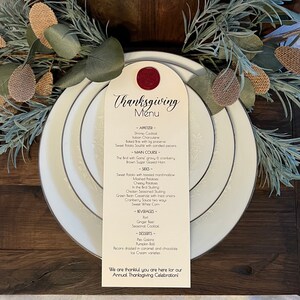 May include: Thanksgiving menu card on a white plate with a silver rim, topped with a red wax seal. The menu lists appetizers, main courses, sides, beverages, and desserts. Decorative greenery and burlap accents surround the plate.