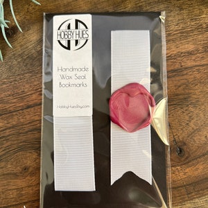 May include: A package containing two white ribbon bookmarks with a burgundy wax seal. The package is black with the text "HOBBY HUES Handmade Wax Seal Bookmarks" and the website address. The bookmarks are packaged in a clear plastic sleeve.