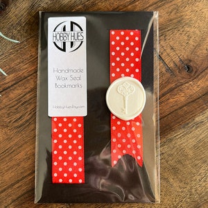 May include: Handmade wax seal bookmarks. The bookmarks feature a red ribbon with white polka dots. A white wax seal with a key design is attached to the ribbon. The package includes a tag with the text "Hobby Hues" and "Handmade Wax Seal Bookmarks".