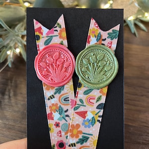 May include: Two decorative bookmarks with floral ribbon and wax seals. One bookmark has a pink wax seal with a tulip design, and the other has a green wax seal with a tulip design. The ribbon has a colourful floral pattern with rainbows.