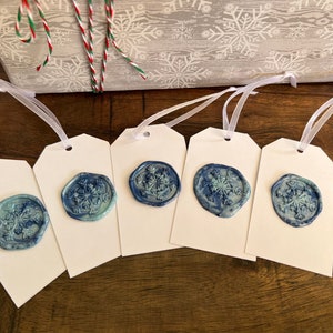 May include: Five white gift tags with blue snowflake wax seals and white ribbon. A wrapped gift with a snowflake pattern and red and green twine is in the background. The tags are on a wooden surface.