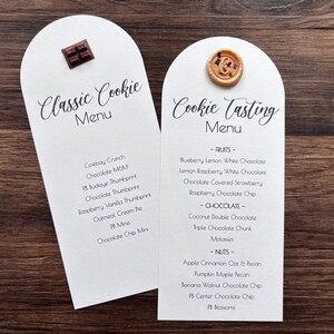May include: Two white arched menu cards with black text. The left card is titled Classic Cookie Menu and features a small chocolate square. The right card is titled Cookie Tasting Menu and features a round tan button with a brown letter G.