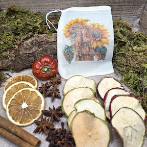 May include: A white drawstring pouch featuring a printed illustration of a person surrounded by sunflowers. The pouch is surrounded by dried orange slices, star anise, whole cloves, dried apple slices, and a small red pumpkin-shaped gourd.