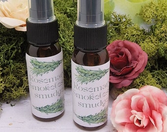 May include: Two small brown spray bottles with black caps and white labels that read "Rosemary Smokeless smudge". The bottles are surrounded by green moss, and pink and red roses.