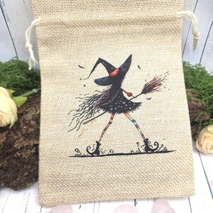Puede incluir: Una bolsa beige de yute con cordón, con una ilustración caprichosa de una bruja con sombrero y vestido negro, sosteniendo una escoba. La bruja tiene pelo largo y suelto y piernas a rayas de colores. La bolsa está decorada con un diseño temático de brujas.